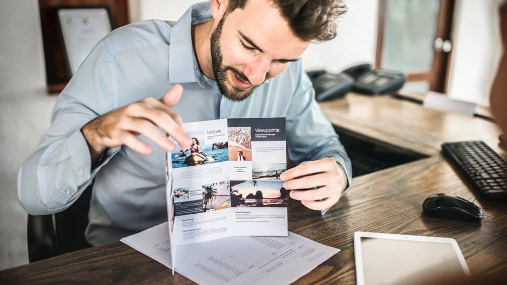 Hombre mostrando un folleto corporativo con fotos y contenido visual mientras conversa en una oficina